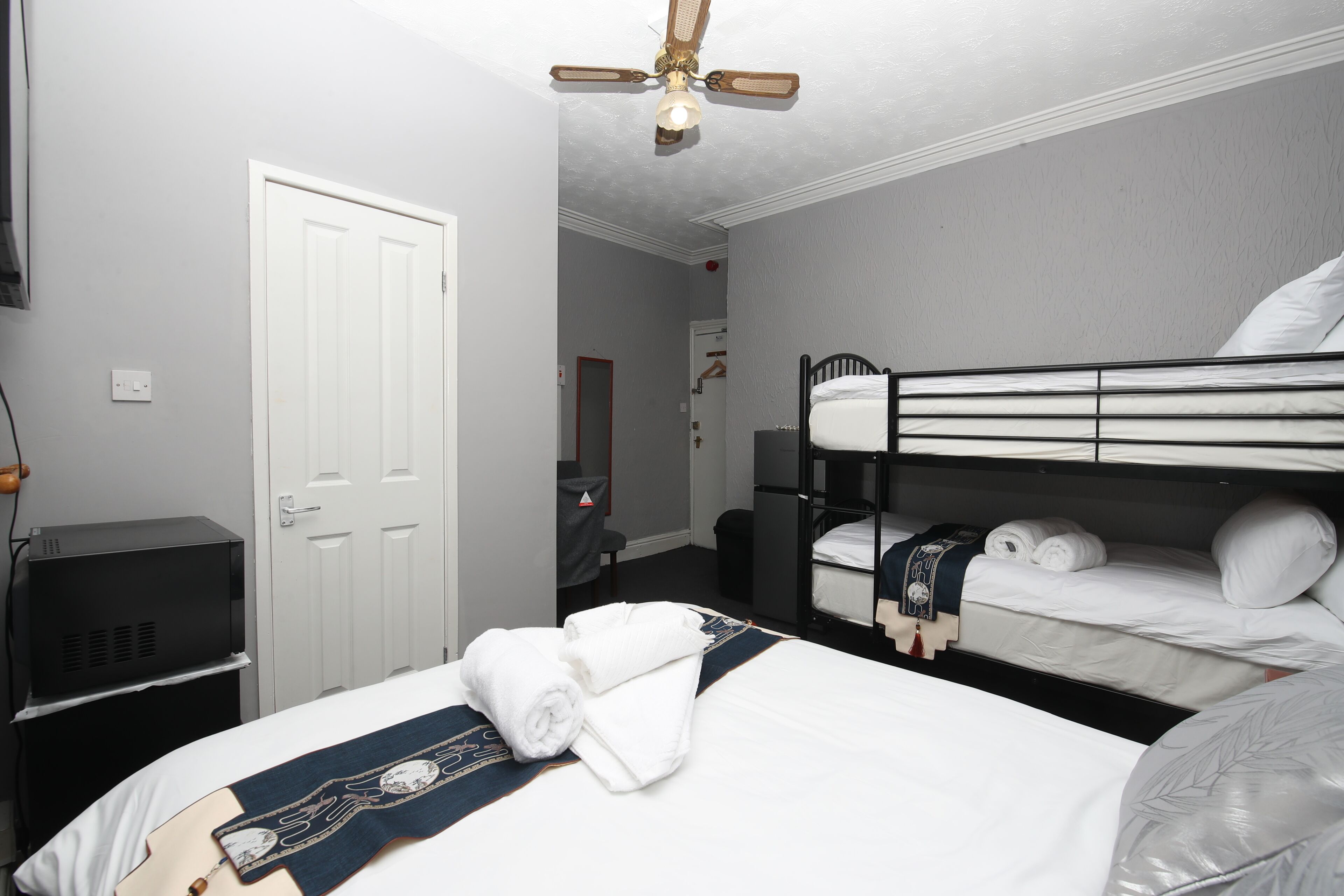 Una habitación de hotel con literas, una cama plana con manta y toallas, un ventilador de techo, un televisor y una puerta.