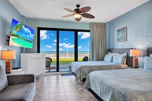 Camera d'albergo con un letto, una TV, una ventola a soffitto, una scrivania, una sedia e vista sulla spiaggia.