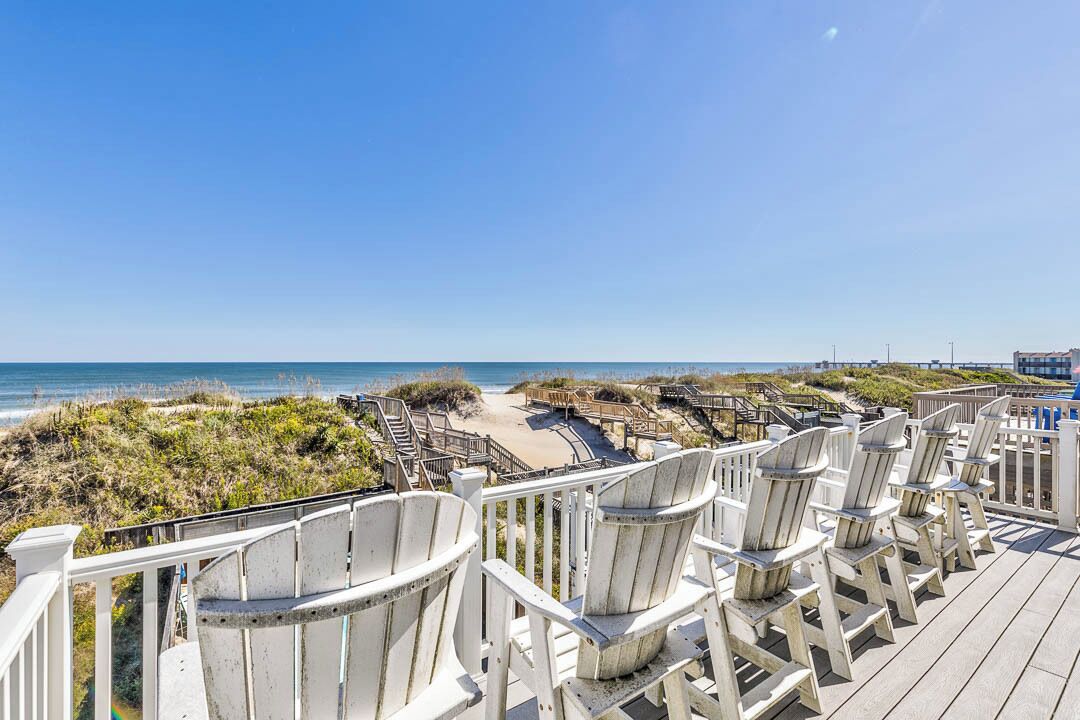 A wooden deck with Adirondack chairs overlooking a beach and boardwalk.