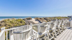 Une terrasse en bois avec des fauteuils Adirondack donnant sur une plage et une promenade en bord de mer.