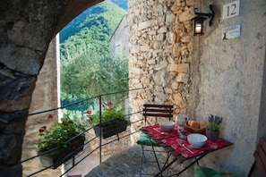 Un muro di pietra rustico con un balcone che si affaccia su una valle boscosa. Un tavolo con tovaglia rossa e diversi oggetti è sistemato contro il muro.
