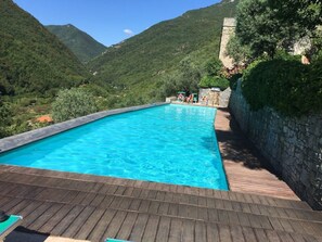 A swimming pool with a wooden deck, surrounded by lush greenery and mountains. - Cassa di Loggia Sleeps 4 With Pool (Castelbianco)
