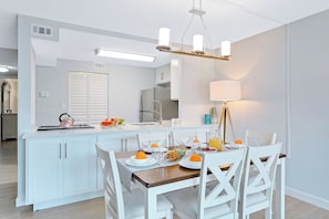 A modern kitchen with white cabinetry, a central island with a kettle, and a dining area with white chairs and a table set for breakfast.