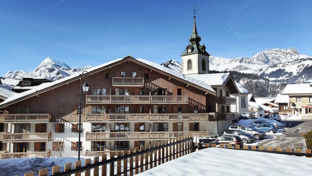 Un bâtiment en bois doté de balcons, une église surmontée d’une flèche, et des montagnes enneigées en arrière-plan.