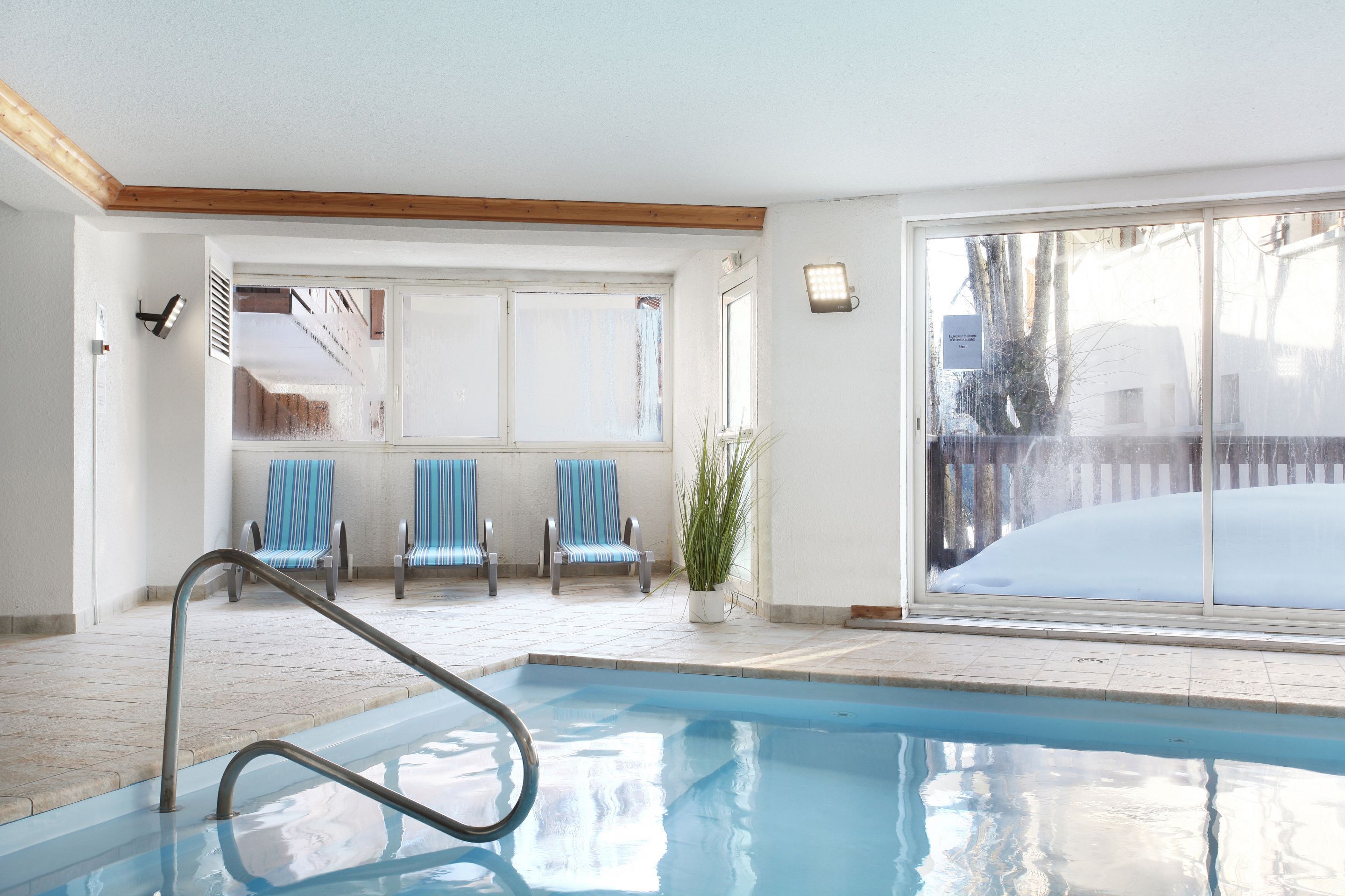 An indoor swimming pool with a metal ladder, blue-striped chairs, and a large window with a view of a snowy landscape.