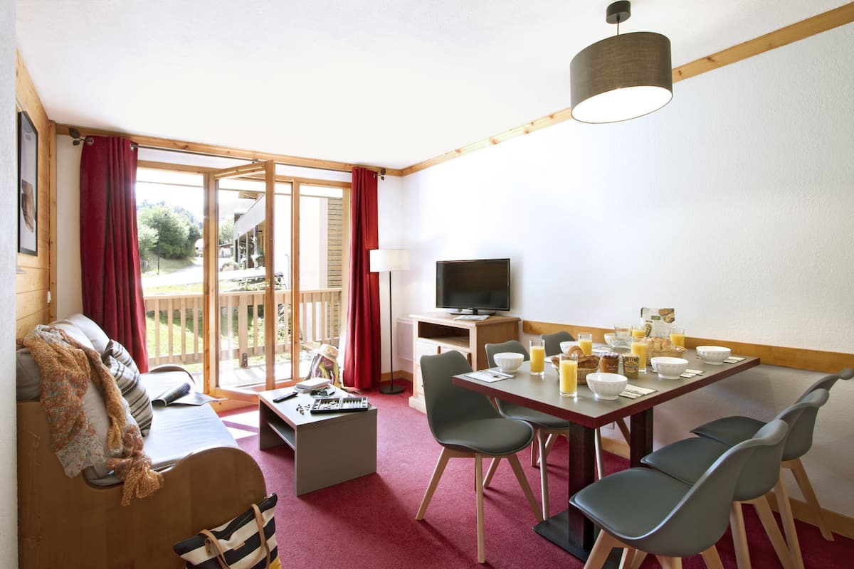 A cozy room with a sofa, a dining table with chairs, a TV, and a window with curtains.