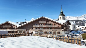 Un immeuble en bois à plusieurs étages, doté de balcons, une église surmontée d’un clocher vert, et des montagnes enneigées en arrière-plan.