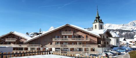 Ein mehrstöckiges Holzgebäude mit Balkonen, eine Kirche mit einem grünen Turm und verschneite Berge im Hintergrund.