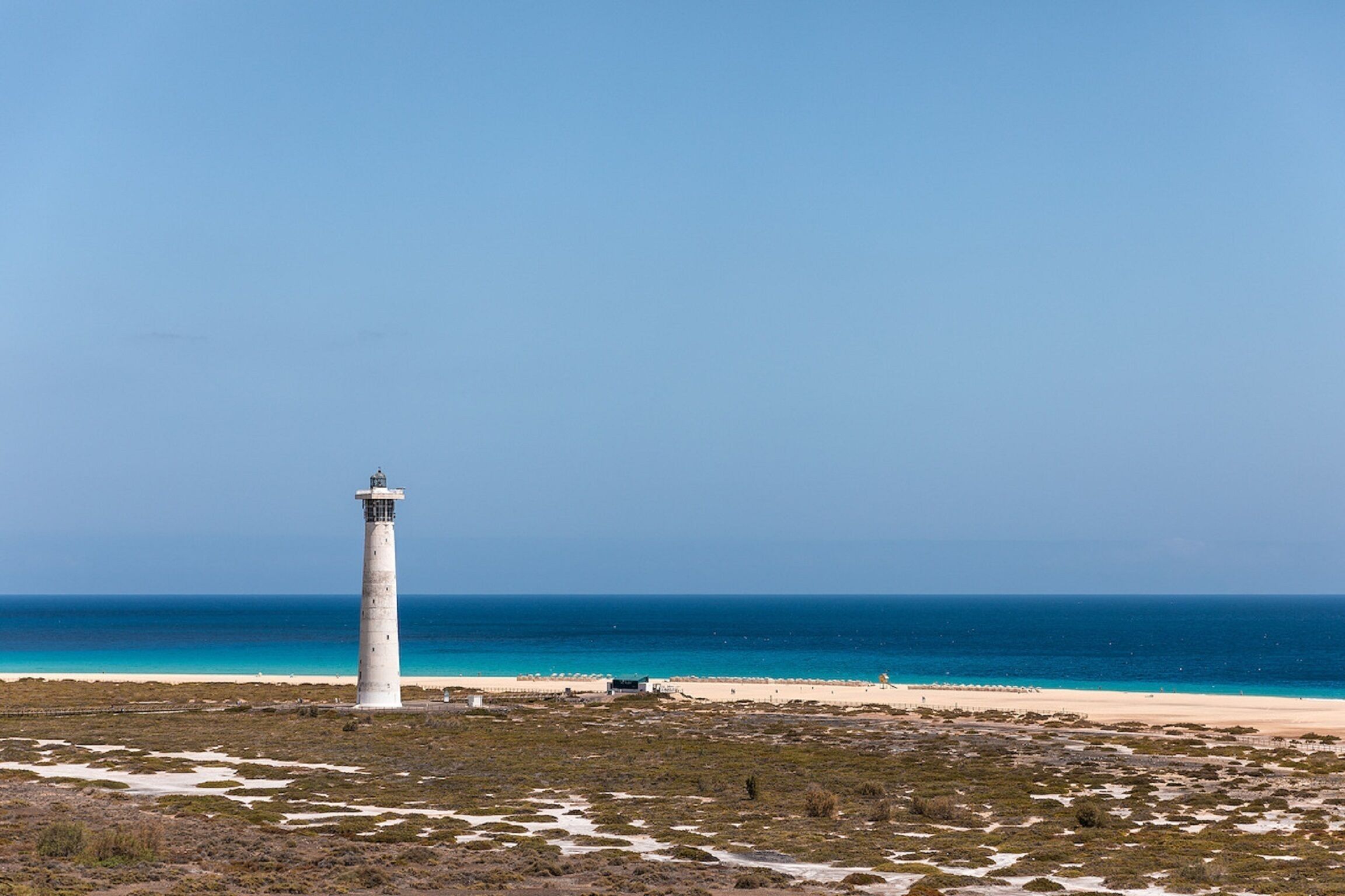 Un phare sur une plage, avec une eau d’un bleu limpide et un ciel dégagé.