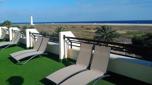 A balcony with green artificial grass and lounge chairs overlooking a beach and a lighthouse. - Apartments Palm Garden in front of the beach (Morro Jable)
