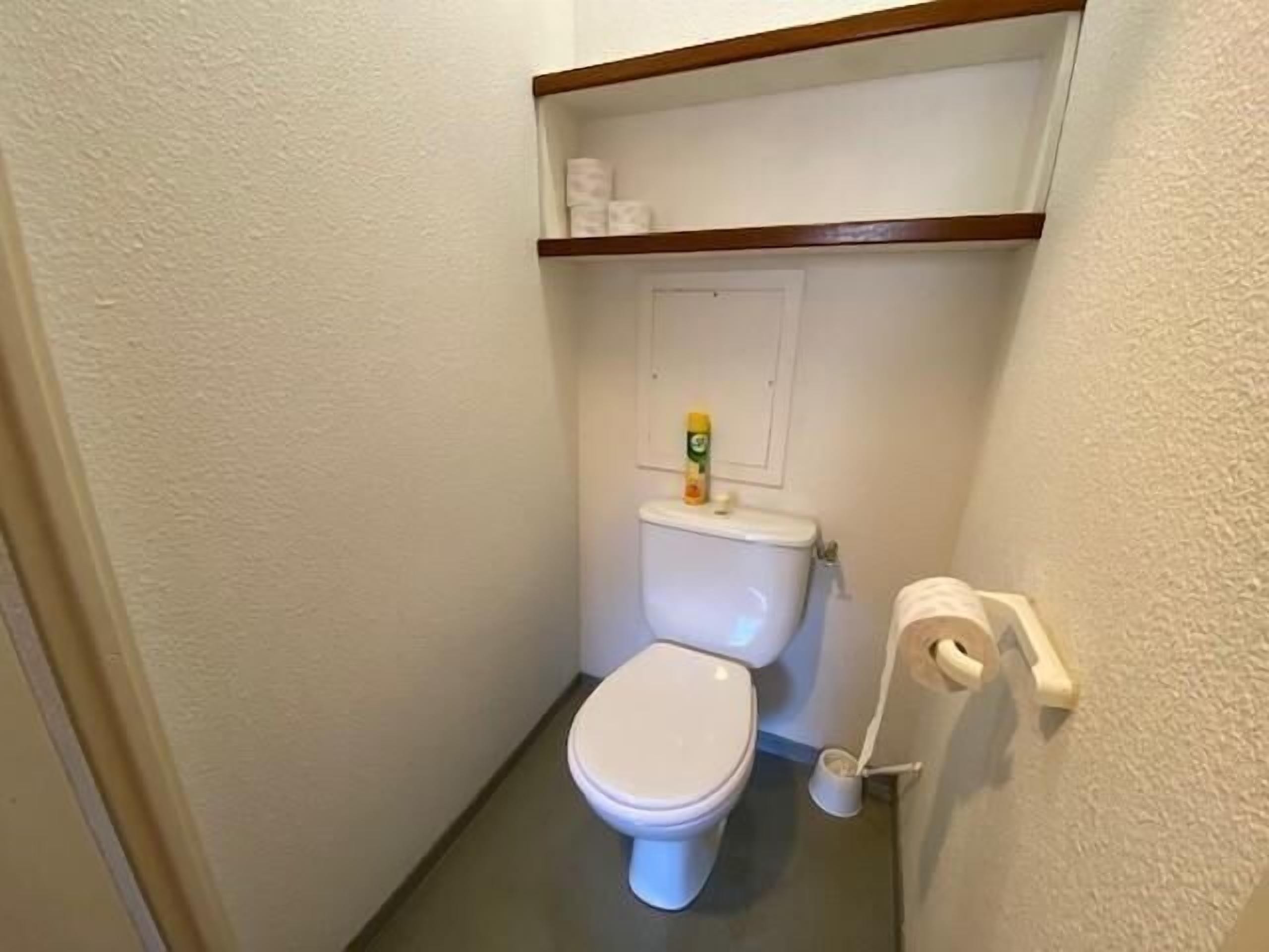 Et lille, kompakt badeværelse med toilet, en hylde over toilettet med toiletpapir og en flaske, samt en toiletbørste på gulvet.