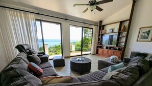 A living room with a large sectional sofa, a round coffee table, and a dog on the sofa. There is a television and a bookshelf in the background. - Stunning views of Lake Arenal & Volcano in Secure Safe Gated Vacation Community (Tronadora)
