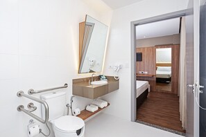 A modern bathroom with a white sink, mirror, and toilet. There are handrails for accessibility, a shower area, and a wooden shelf with towels. - SLAVIERO Curitiba Aeroporto (Sao Jose dos Pinhais)
