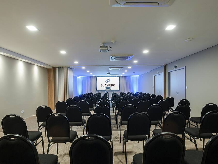 A conference room with rows of black chairs, a projector screen, and a sign reading "SLAVIERO".
