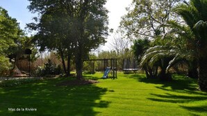 A well-maintained grassy area with a playground featuring a slide and swings, surrounded by lush trees and palm plants.