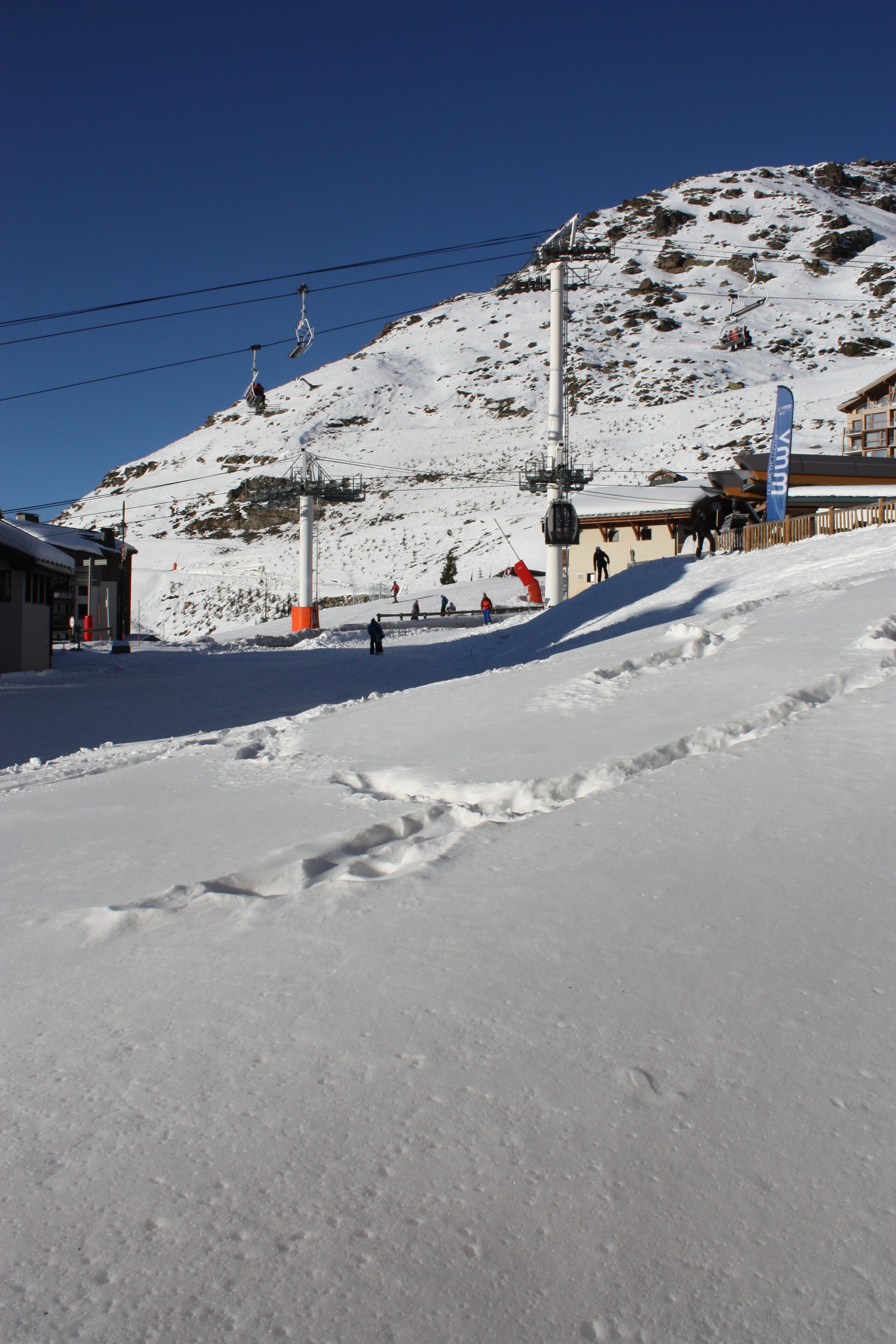 A snow-covered ski slope with a ski lift, buildings, and a clear blue sky.