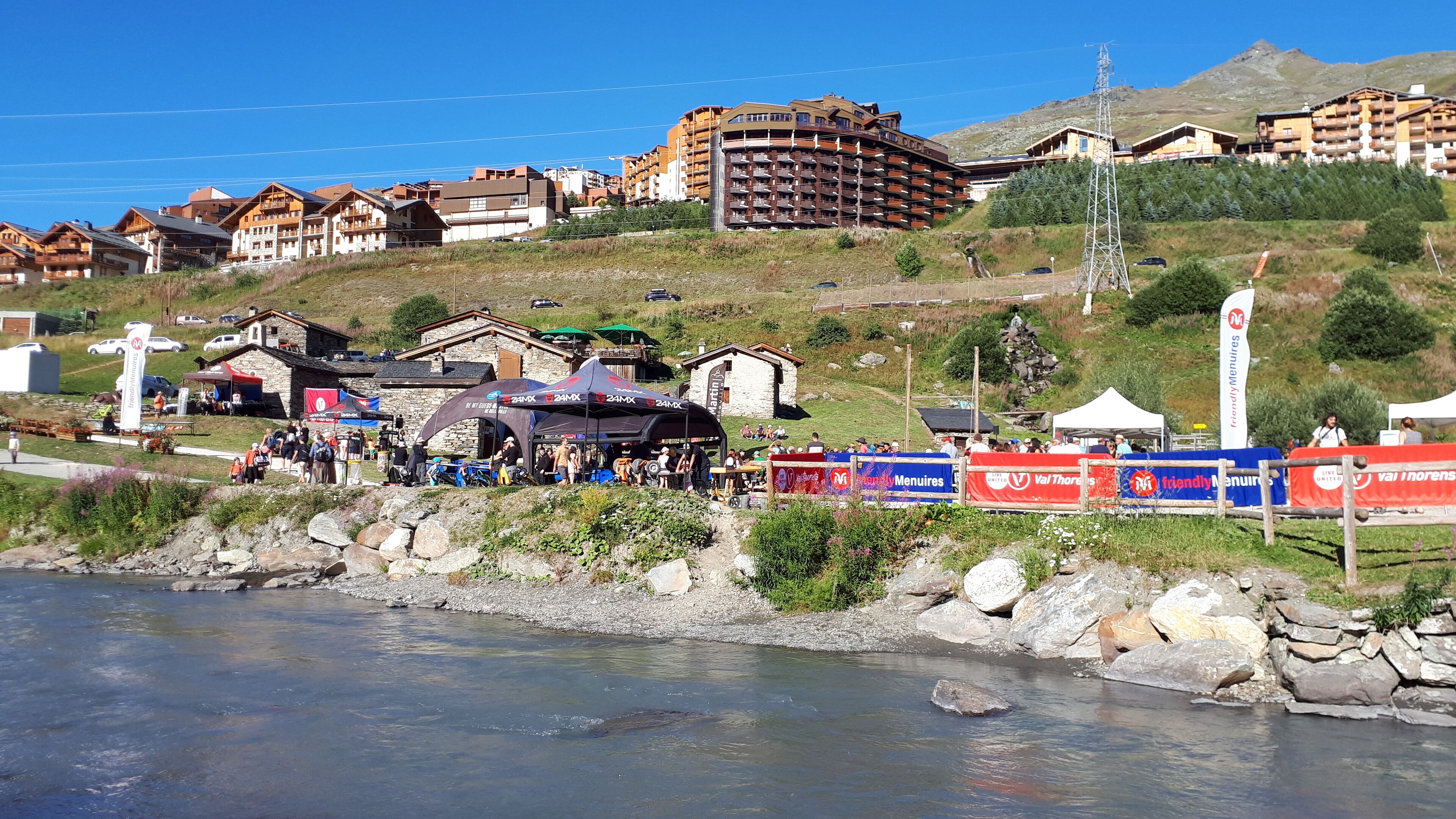 A riverside view of a mountain resort with multiple lodging buildings, a clear sky, and a rocky shoreline.