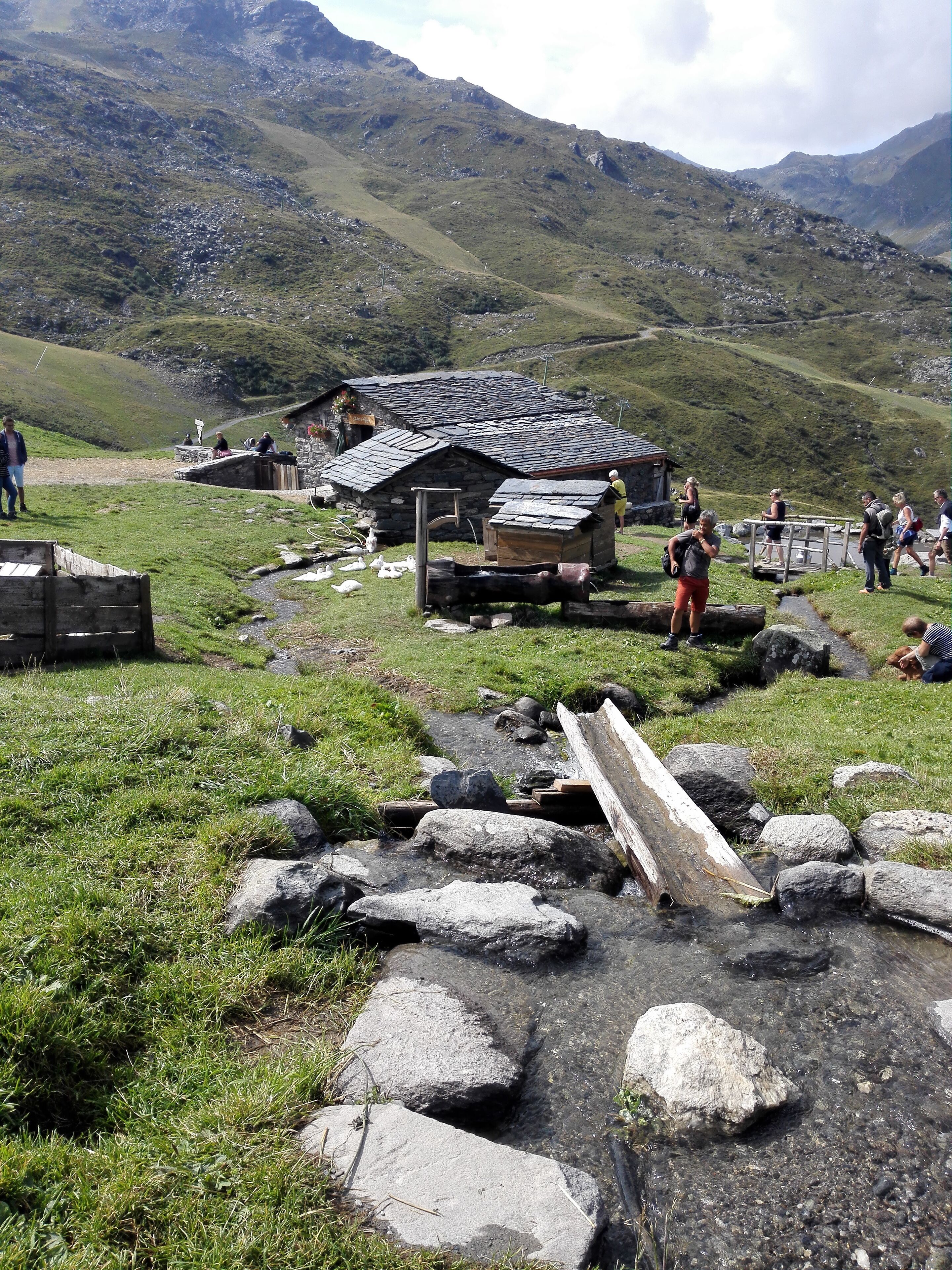 Un bâtiment rustique en pierre, doté d’un toit en pente, entouré d’une zone herbeuse parsemée de rochers.