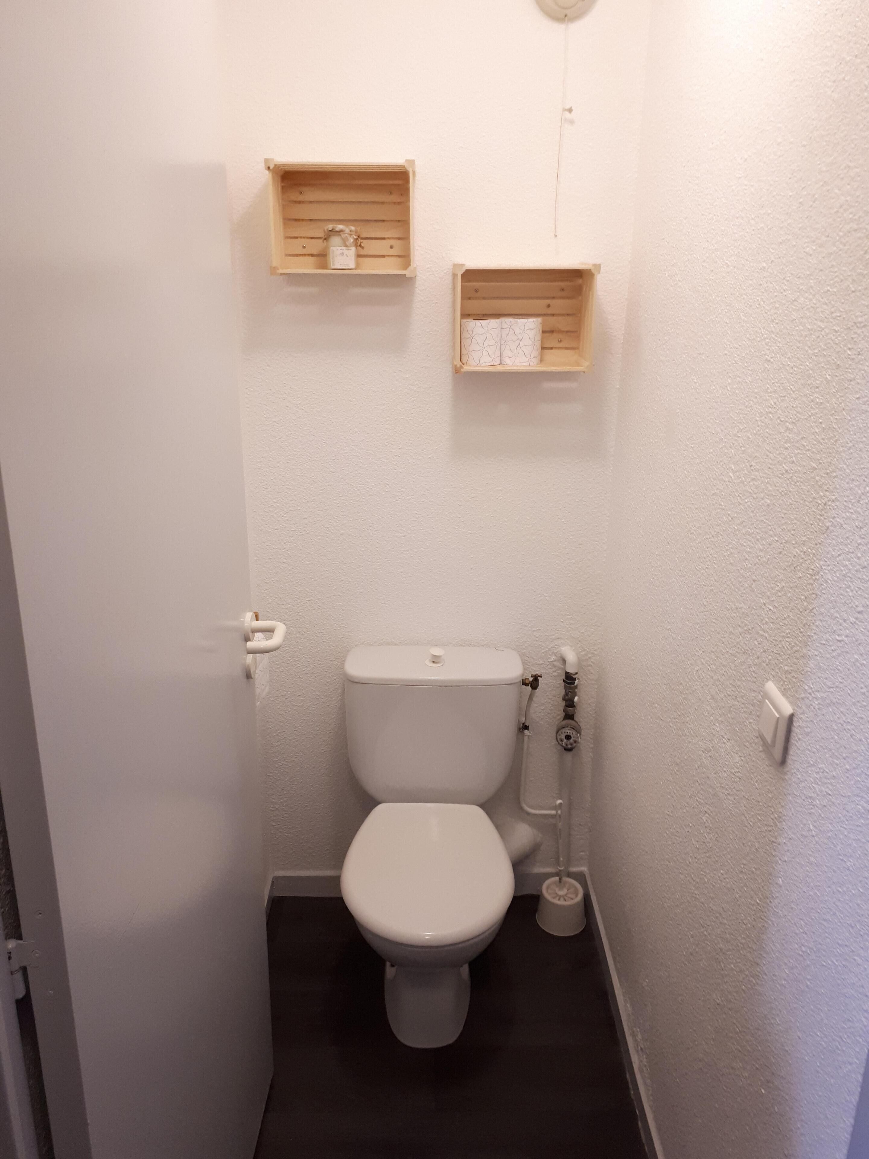 Ein kleines, schmales Badezimmer mit einer weißen Toilette, einem Holzregal darüber und einem Toilettenpapierhalter an der Wand.