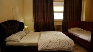 A bedroom with a large bed, a smaller bed, a nightstand, a lamp, and a window with curtains. - Moon Hostel (Pristina)