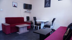 A small, modern room with a red sofa, a white table, and a black chair. There is a flat-screen TV mounted on the wall and a small shelf below it. - Moon Hostel (Pristina)