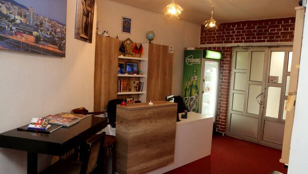 A reception area with a wooden reception desk, a computer, and a bookshelf with various items. There is a globe on the wall and a sign for Tuborg beer. - Moon Hostel (Pristina)