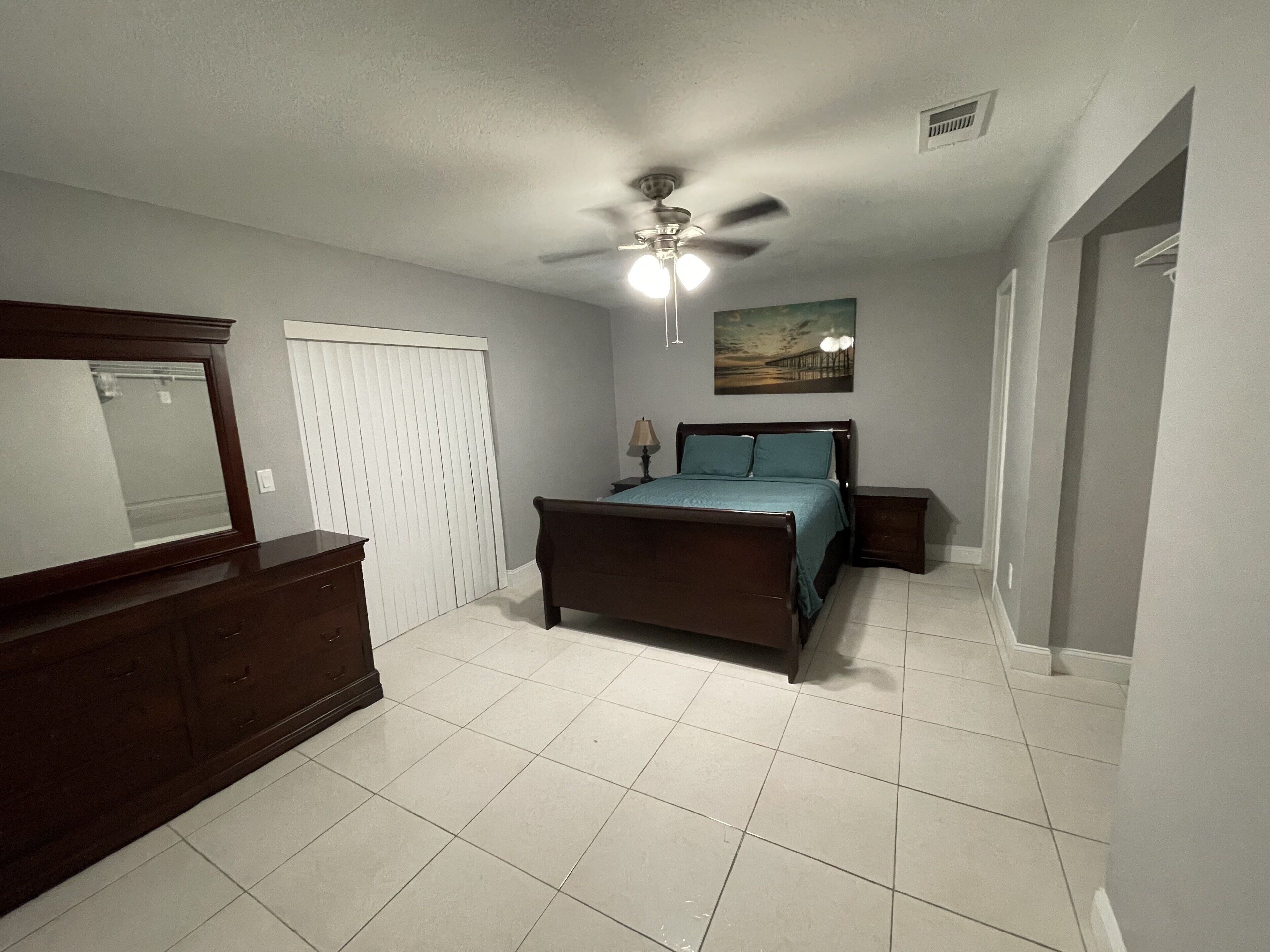 A bedroom with a bed, dresser, ceiling fan, and a painting on the wall.