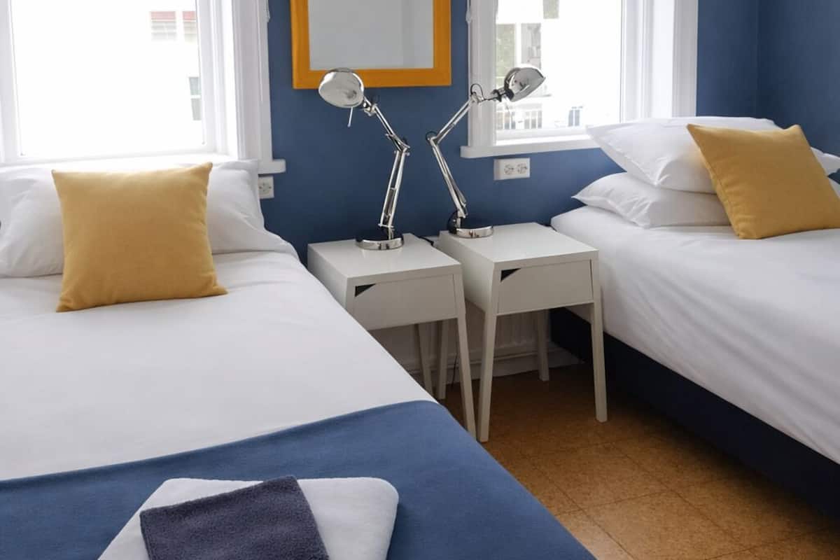 Habitación de hotel con dos camas, ropa de cama blanca, almohadas amarillas, un escritorio con lámpara y un cuadro en la pared.