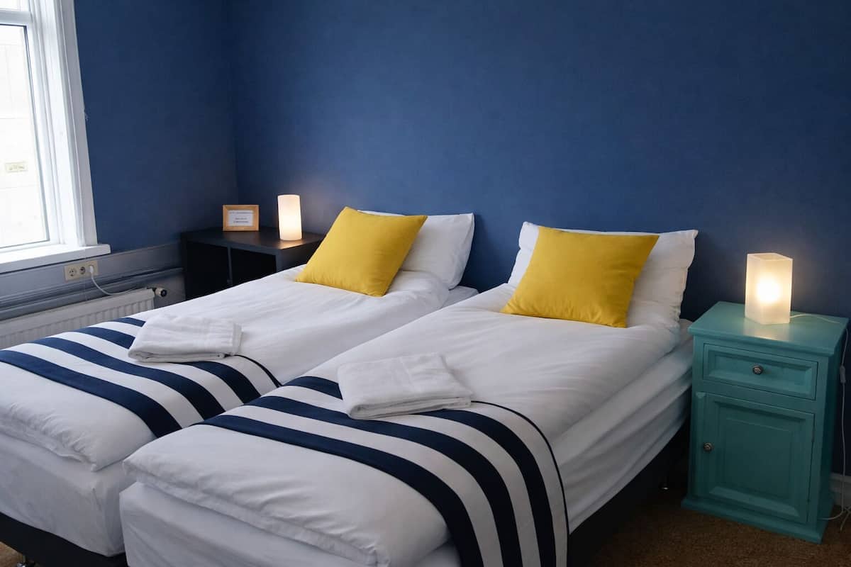 Una habitación de hotel con dos camas, cada una con ropa blanca y almohadas a rayas azules. Hay mesitas de noche con lámparas y una mesita auxiliar con una lámpara.