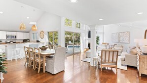 A spacious living area with a dining table, chairs, and a sofa. There is a kitchen area with white cabinets and a large window. - 4 Scaup Court:  A BESTNEST by Beverly Serral (Hilton Head Island)