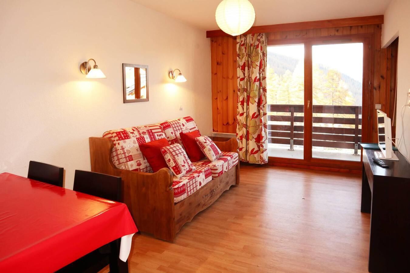 Um quarto aconchegante com piso de madeira, uma mesa vermelha, um sofá de madeira com almofadas estampadas e uma janela grande com cortinas.