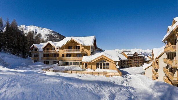Un chalet enneigé composé de plusieurs bâtiments en bois, avec une piste de ski et des montagnes en arrière-plan.