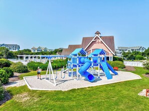A playground with slides and swings, surrounded by a well-maintained lawn and a building with a brown roof in the background.