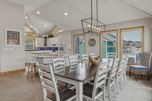A spacious kitchen with white cabinets, a dining area with a large wooden table and white chairs, and a view of a blue house through a window. - Duck Tales | 2400 ft from the beach | Private Pool, Hot Tub | Duck (Duck)