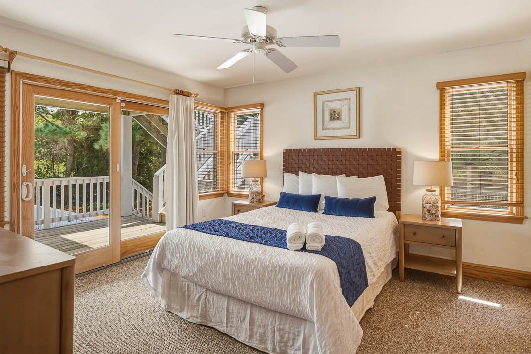 A bedroom with a large bed, a ceiling fan, a window with blinds, and a view of a deck.