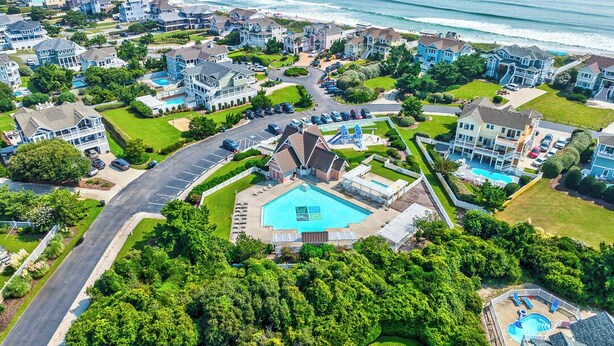 An aerial view of a coastal residential area with houses, a central swimming pool, and a road. - Duck Tales | 2400 ft from the beach | Private Pool, Hot Tub | Duck (Duck)