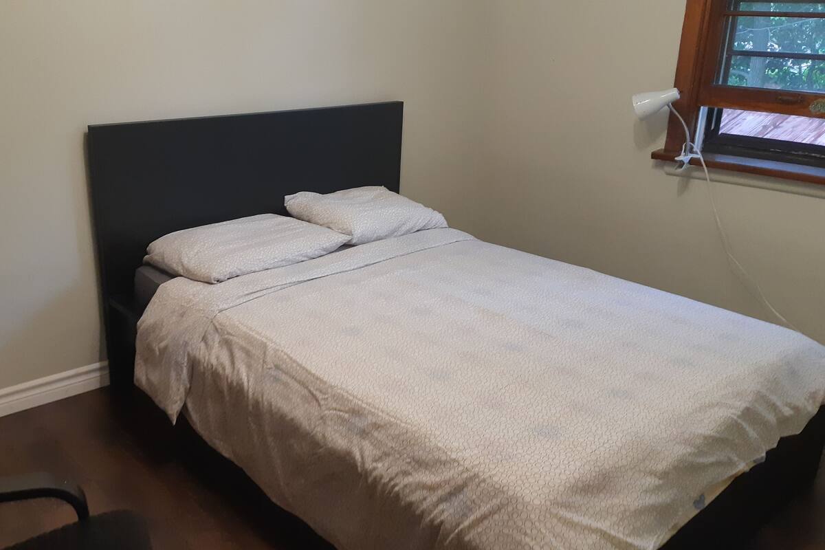 A small, neatly made bed with a dark headboard and white bedding in a room with a wooden dresser and a window with a view of trees.