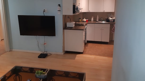 Un espacio habitable compacto con un televisor de pantalla plana montado en la pared, una pequeña kitchenette con pileta y alacenas, y un comedor con mesa y sillas.