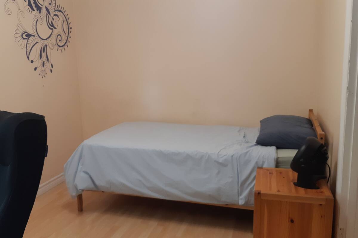 A small, single-bed room with a wooden nightstand, a chair, and a wall decal.