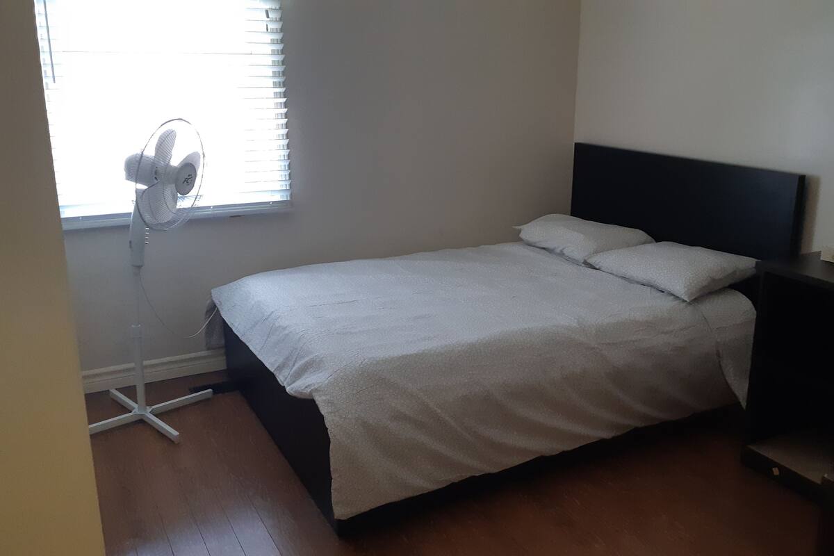 A bedroom with a bed, a standing fan, and a window with blinds.
