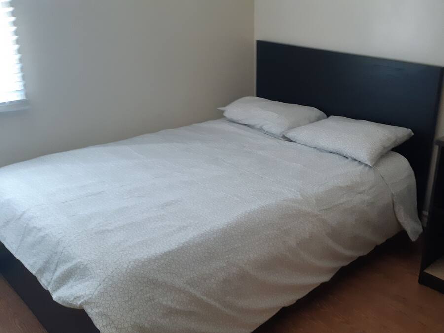 Una cama individual con edredón blanco y dos almohadas, cabecera oscura y mesita de noche con una caja encima.