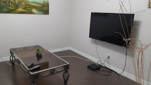 Una sala de estar con un televisor de pantalla plana montado en la pared, una mesa de centro de vidrio con un control remoto y una pequeña planta.