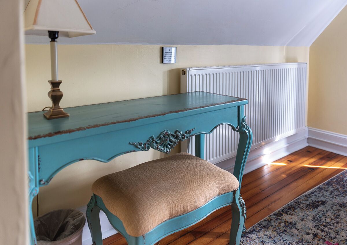 Una silla color turquesa con asiento de arpillera en una habitación con suelo de madera, un radiador y una lámpara.