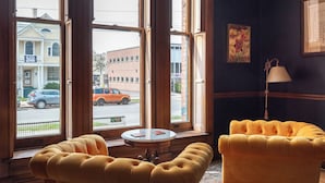 Une pièce avec deux fauteuils jaunes à motifs capitonnés, une petite table ronde et une lampe. De grandes fenêtres offrent une vue sur la rue et les bâtiments environnants.