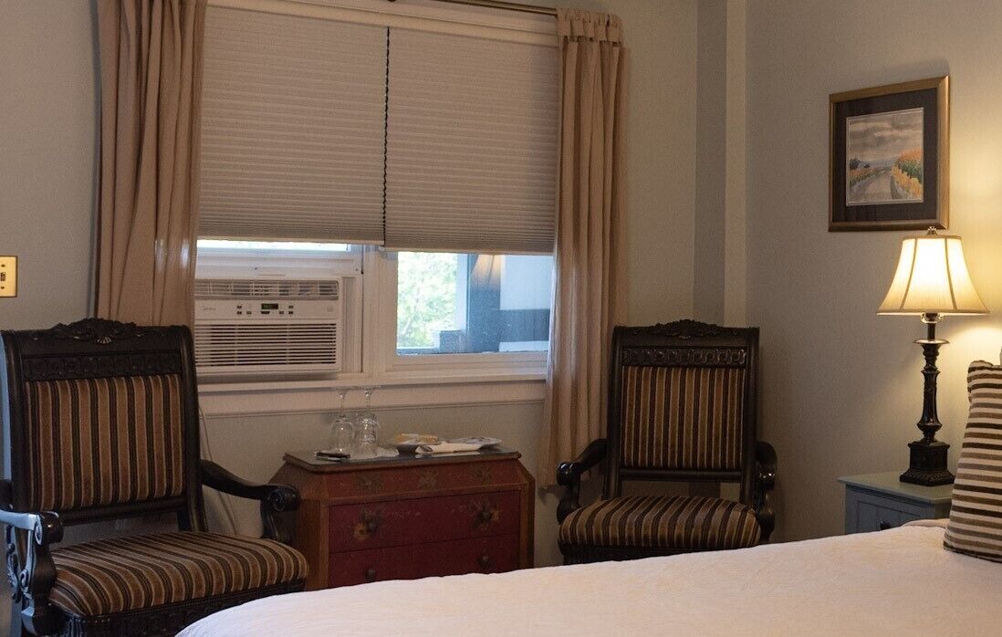 A hotel room with a bed, two upholstered chairs, a wooden nightstand, a lamp, and a window with curtains.