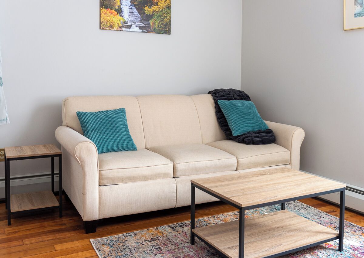 Un sofá color beige con cojines verde azulado y negros, una mesita de madera y una mesa de centro con un tapete colorido.