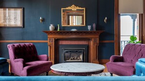 Une pièce avec une cheminée, deux fauteuils violets, une table ronde et un miroir au-dessus de la cheminée.