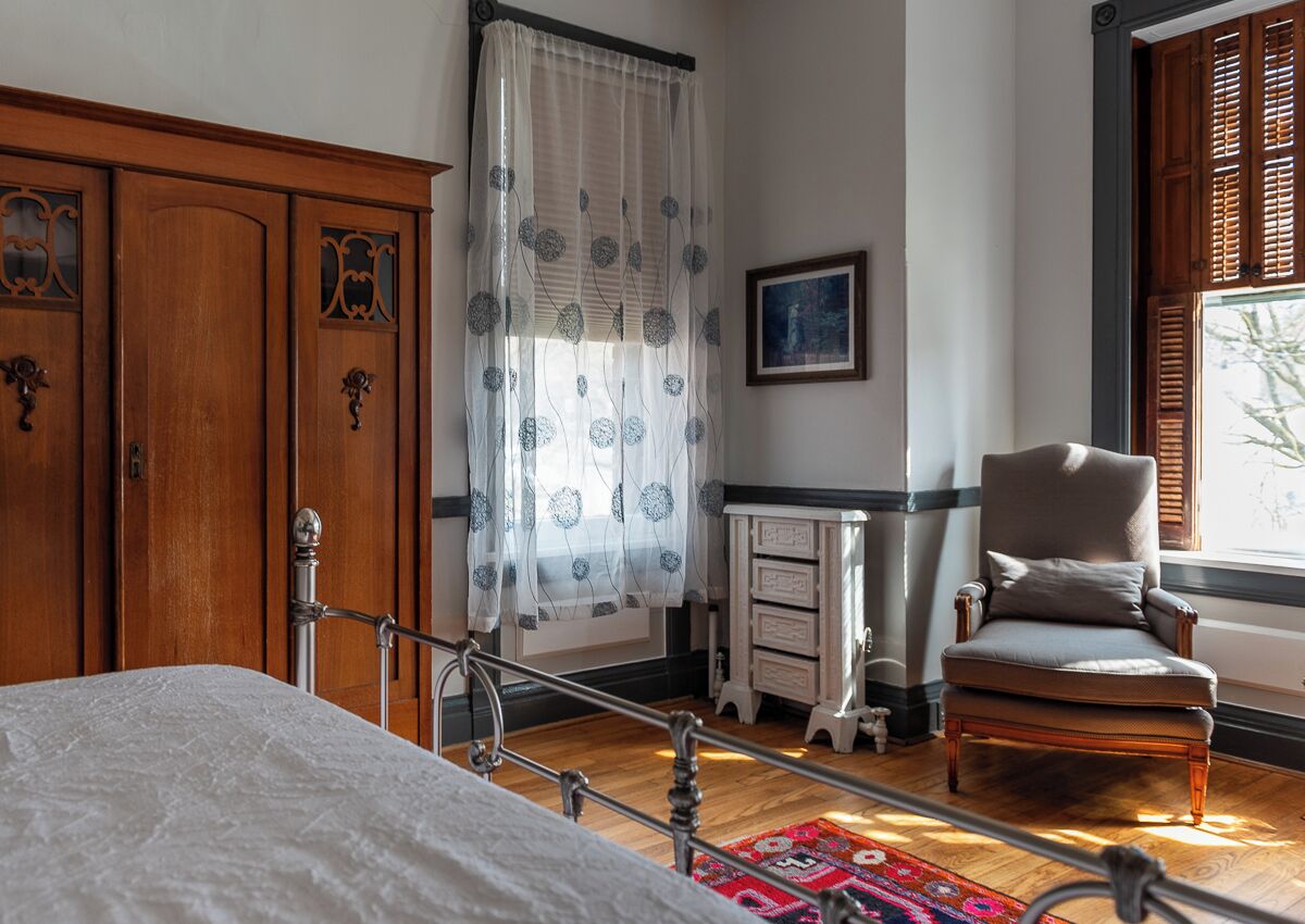 A bedroom with a bed, a wooden wardrobe, a chair, a small table, and a window with shutters.