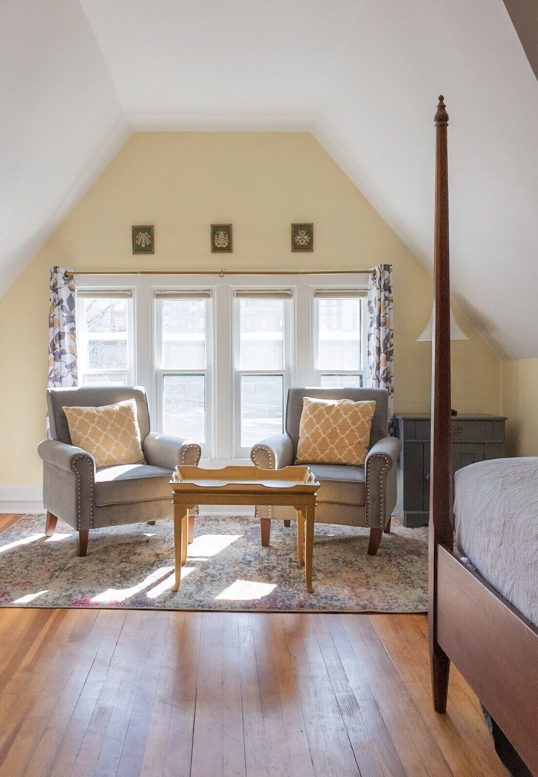 A bedroom with a sloped ceiling, two armchairs, a wooden coffee table, a bed, and a dresser.