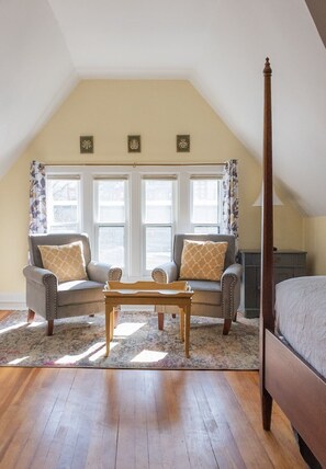 A bedroom with a sloped ceiling, two armchairs, a wooden coffee table, a bed, and a dresser.